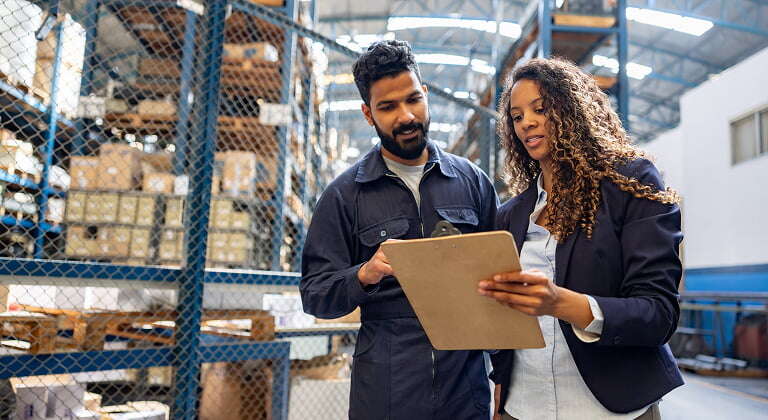 Manager talking to employee in industrial warehouse showing him inventory on clipboard